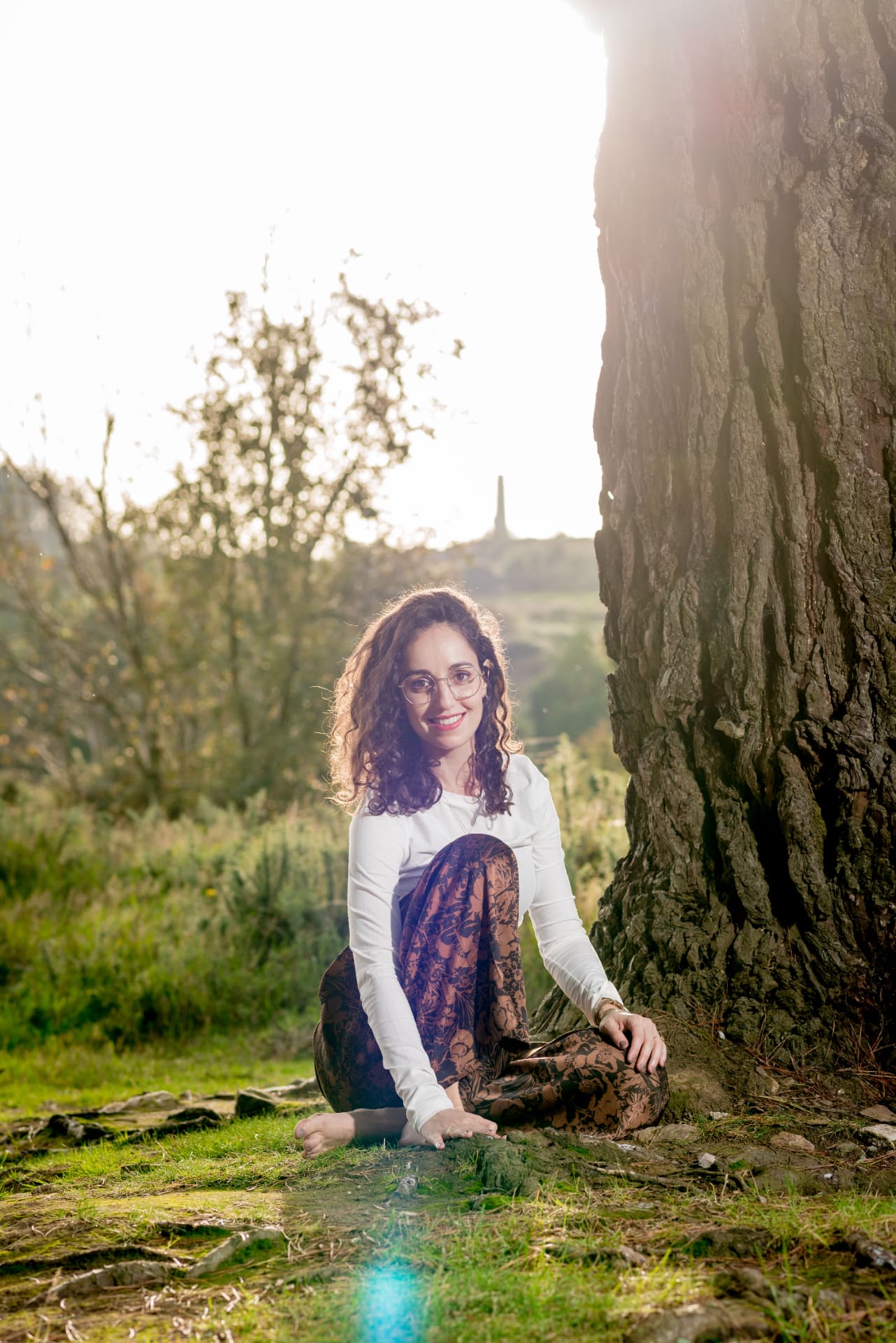 Roni sitting barefoot beside a large tree, smiling warmly